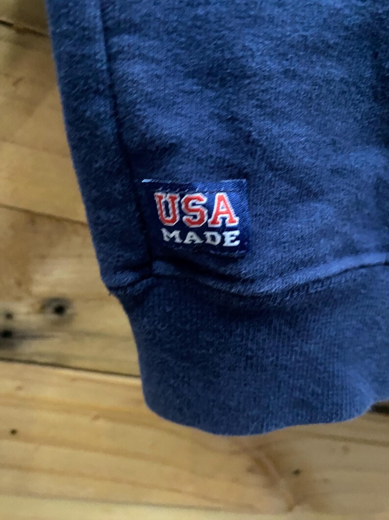 May include: Close-up of a navy blue garment with a ribbed cuff. A rectangular patch with the text "USA MADE" in red and white is sewn onto the garment. The background is a wooden surface.