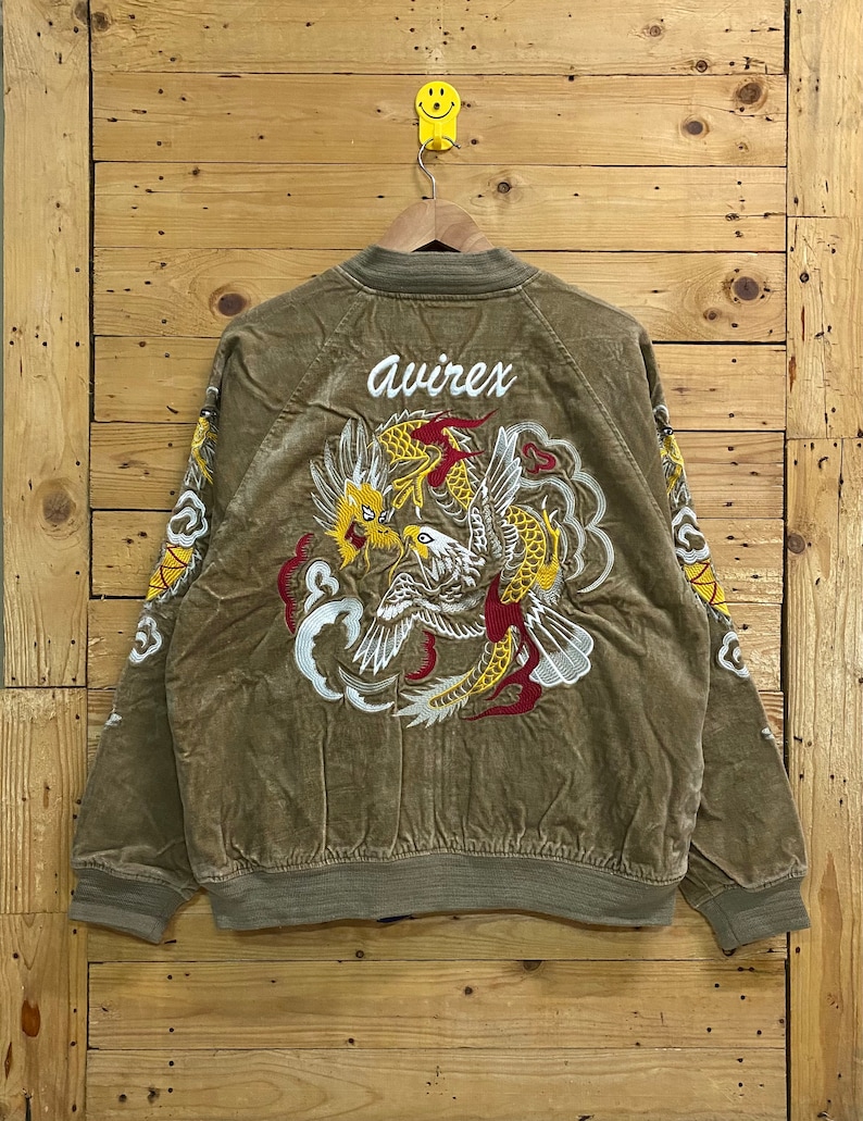 Pu&ograve; includere: Una giacca bomber marrone con un grande disegno ricamato di un drago e un uccello sul retro. La giacca ha la parola "avirex" ricamata sul retro.
