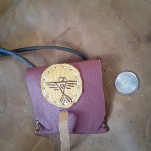 May include: A small, burgundy leather pouch with a flap closure and a yellow leather circle with a brown bird design in the center. The pouch has a brown leather strap and is hanging from a black cord.
