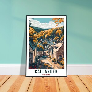Op de afbeelding: Een ingelijste kunstprint met een gestileerde illustratie van Callander, Schotland. Het kunstwerk toont een straatbeeld met gebouwen en een weg, omgeven door herfstkleurige bomen en een blauwe lucht. De tekst "CALLANDER SCOTLAND" staat onderaan.