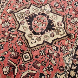 May include: Close-up of a vintage rug with a detailed floral pattern. The rug features a central medallion in cream, black, and pink, surrounded by a red field with intricate designs. The rug's border is dark brown.
