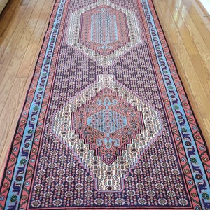 May include: A long, rectangular rug with a traditional Persian design. The rug has a dark blue background with intricate patterns in shades of pink, red, blue, and white. The rug has a fringed border.
