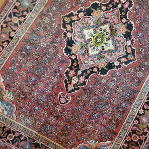 Puede incluir: Una alfombra persa vintage con fondo rojo y intrincados patrones florales en tonos de rosa, azul, verde y negro. La alfombra tiene un medallón central con un diseño de estrella y un borde con un patrón geométrico repetitivo.