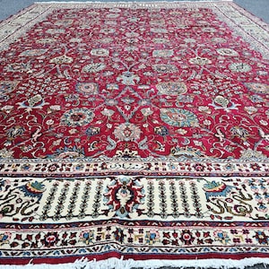 May include: A large, rectangular red rug with an intricate floral pattern. The rug features a cream-coloured border with detailed designs and a fringed edge. The central field is filled with a dense arrangement of flowers and leaves in shades of blue, green, and cream.