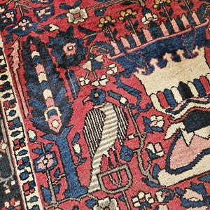 May include: A woven rug featuring a vibrant red background with intricate floral patterns in blue, white, and yellow. The design includes a central bird figure with striped wings, stylised trees, and geometric borders, showcasing a detailed textile weave.