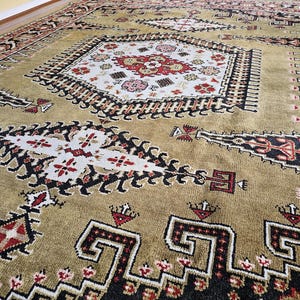 Könnte beinhalten: Ein aufwendig gemusterter Teppich mit einem zentralen Medaillon-Design. Der Teppich hat einen beigefarbenen Hintergrund mit schwarzen, roten und weißen geometrischen Mustern. Der Rand des Teppichs ist dunkler und setzt einen Kontrast.