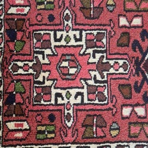 May include: Close-up of a section of a red oriental rug with a detailed geometric pattern. The design features white, brown, and black shapes, creating a symmetrical and intricate visual texture. The rug's border has a dark blue and brown design.