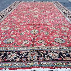 May include: A large, rectangular red rug with an intricate floral pattern. The rug features a central field of red with blue, cream, and gold floral motifs. A black border with detailed designs frames the rug, and white fringe edges the bottom.