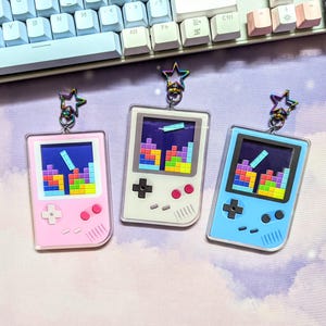 以下が含まれることがあります： レトロな携帯ゲーム機の形をしたカラフルなキーホルダーチャーム3つ。各チャームには、ピクセル化されたテトリスのゲーム画面とカラフルなボタンのデザインが施されています。チャームはピンク、白、青です。