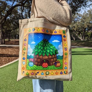 May include: Beige canvas tote bag with a vibrant illustration of a treehouse with a green leafy roof, blue sky, and various fruits and vegetables. The bag has a rectangular design with a border of colourful icons.