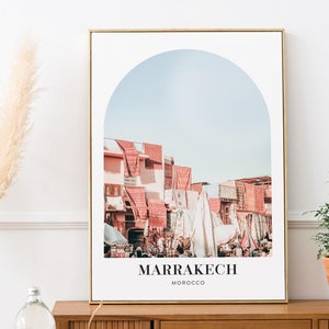 May include: A framed print with a photograph of a city street scene in Marrakech, Morocco. The image is framed in a gold frame and features a rounded archway with a blue sky in the background. The text "MARRAKECH" and "MOROCCO" is printed below the image.