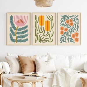 Peut inclure: Trois impressions d'art botanique encadrées. Les impressions présentent des motifs floraux abstraits colorés en rose, orange et turquoise sur un fond crème. L'œuvre est exposée au-dessus d'un canapé blanc avec des coussins aux tons neutres.