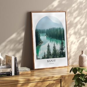 May include: A framed print of a scenic landscape with a lake surrounded by trees and mountains. The print has a rounded arch shape and the text "BANFF CANADA" at the bottom.
