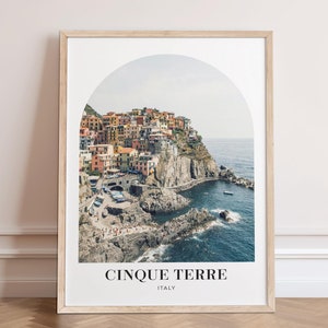 May include: A framed print of a colourful Italian village perched on a cliff overlooking the ocean. The image is framed in a natural wood frame and features an arched window shape. The text "Cinque Terre, Italy" is printed below the image.