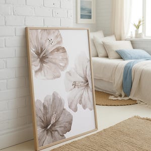 May include: A framed art print featuring watercolor hibiscus flowers in shades of taupe and white. The artwork is displayed in a light wood frame and leans against a white brick wall. The room has a neutral color palette with a woven rug.
