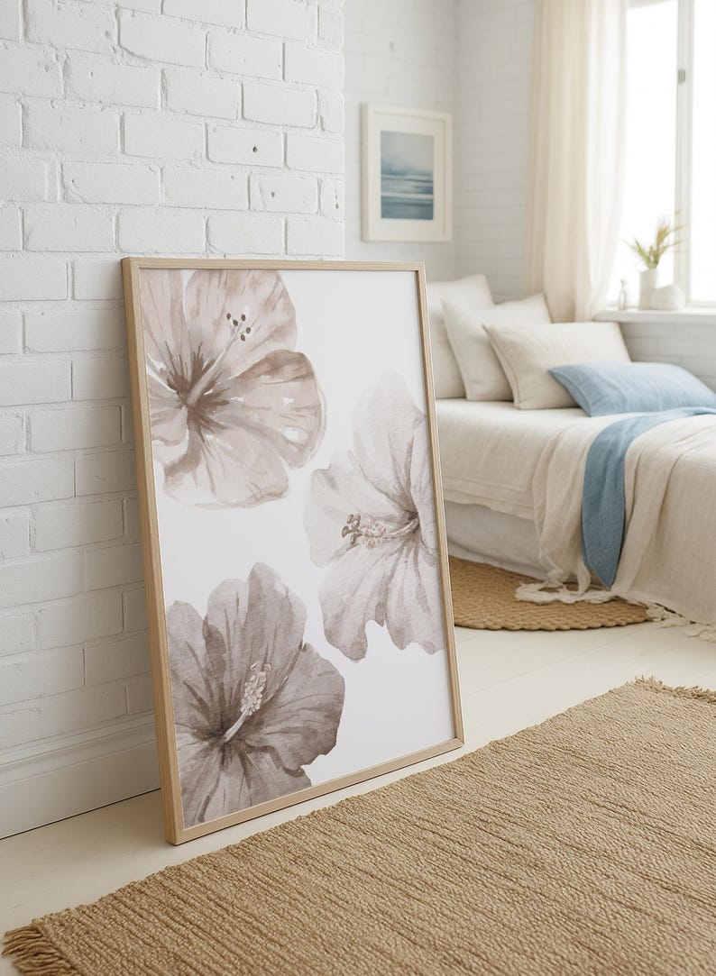 May include: A framed art print featuring watercolor hibiscus flowers in shades of taupe and white. The artwork is displayed in a light wood frame and leans against a white brick wall. The room has a neutral color palette with a woven rug.