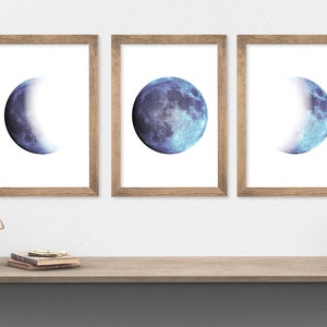 May include: Three framed prints of a blue watercolor moon in different phases. The moon is in a crescent, half-moon, and full moon phase. The prints are in simple wooden frames.