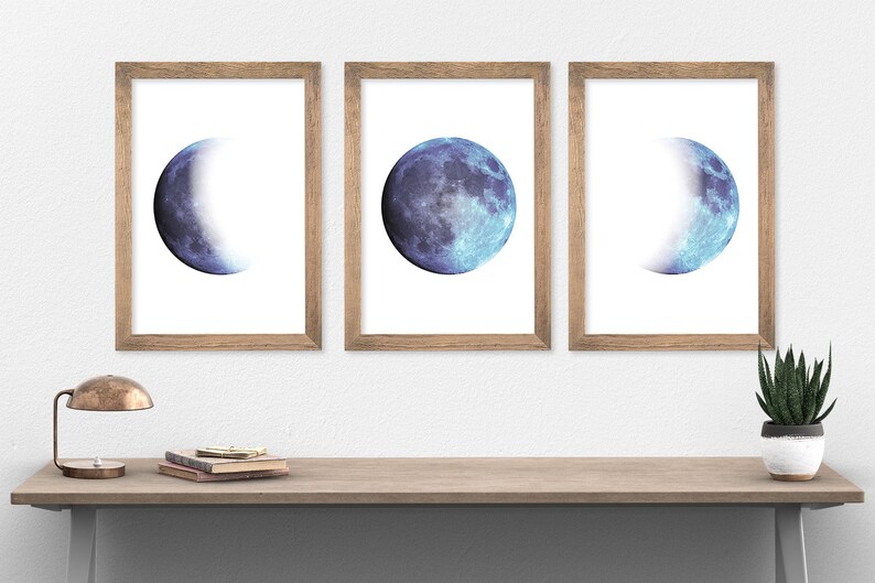 May include: Three framed prints of a blue watercolor moon in different phases. The moon is in a crescent, half-moon, and full moon phase. The prints are in simple wooden frames.