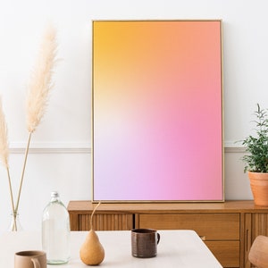 Puede incluir: Una impresión de arte abstracto enmarcada con un degradado de colores amarillo, naranja, rosa y morado. La impresión está colgada en una pared blanca sobre una cómoda de madera con un jarrón de hierba de la pampa seca y una planta en maceta.