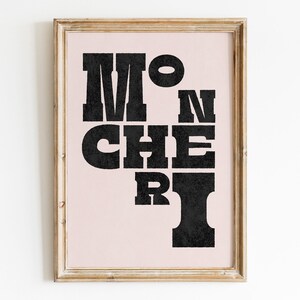 May include: A framed print with a pink background and black text that reads "Mon Cheri". The text is in a bold, blocky font.