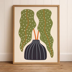May include: A framed art print featuring a stylized illustration of a plant. The artwork showcases two large, green leaves with white polka dots, orange stems, and a black vase with white vertical lines. The background is a light beige.