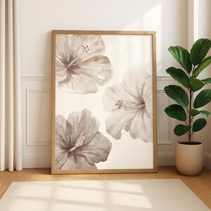 May include: A framed floral art print featuring three large hibiscus flowers in shades of gray and beige. The artwork is displayed in a light wood frame and leans against a white wall. A potted plant sits to the right.