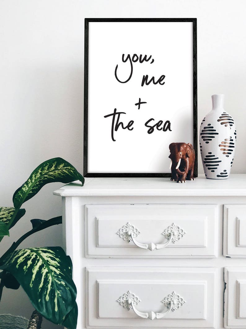 May include: A black framed print with the text "you, me + the sea" in white cursive writing. The print is sitting on a white dresser with two drawers.