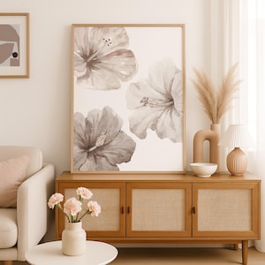 May include: A framed floral print featuring three large hibiscus flowers in shades of gray and white. The artwork is displayed above a wooden cabinet with woven cane door panels. A vase of pampas grass and a small lamp are on the cabinet.