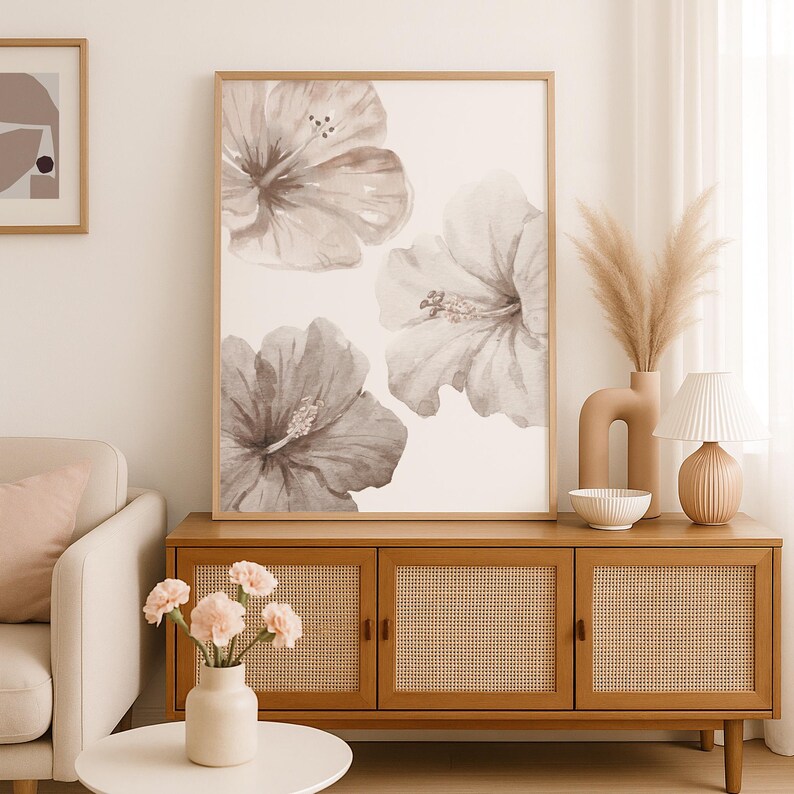 May include: A framed floral print featuring three large hibiscus flowers in shades of gray and white. The artwork is displayed above a wooden cabinet with woven cane door panels. A vase of pampas grass and a small lamp are on the cabinet.