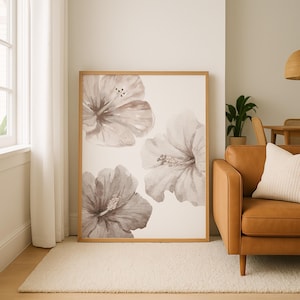 May include: A framed art print featuring three watercolor hibiscus flowers in shades of gray and brown. The artwork has a light background and is displayed in a natural wood frame. The print is leaning against a wall in a living room setting.