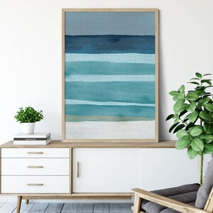 May include: A framed abstract watercolor painting with horizontal blue and teal stripes, representing the ocean and sky. The painting is displayed on a white dresser with a wooden top, a gray armchair, and a potted plant.