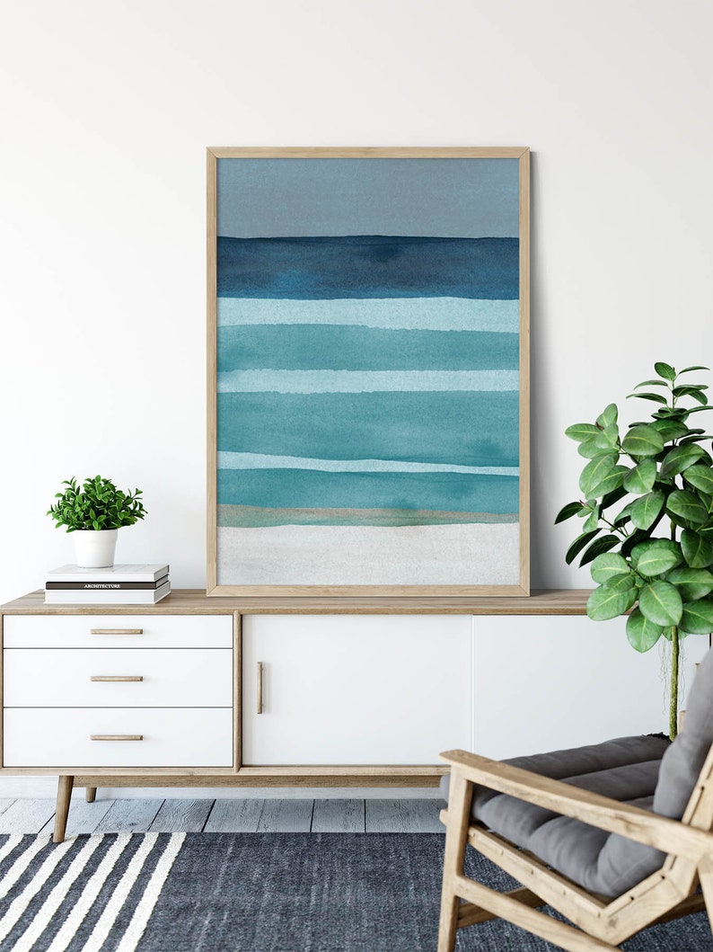 May include: A framed abstract watercolor painting with horizontal blue and teal stripes, representing the ocean and sky. The painting is displayed on a white dresser with a wooden top, a gray armchair, and a potted plant.