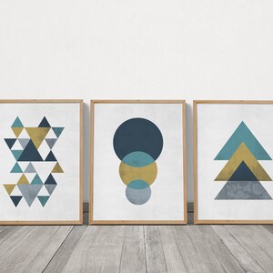 以下が含まれることがあります： 青、ターコイズ、ゴールドの色の抽象的な幾何学模様が描かれた、額装されたプリント3枚セット。プリントには、三角形の模様、円の模様、三角形の積み重ね模様が描かれています。