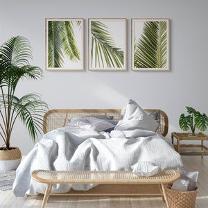 May include: A white bed with a light wood frame and a white linen duvet. The bed is decorated with pillows and a throw blanket. There are three framed prints of palm leaves above the bed. A small wooden side table with a potted plant sits next to the bed. A wicker bench sits at the foot of the bed.