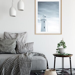Pu&ograve; includere: Un faro bianco si erge alto contro un cielo nuvoloso, incorniciato da una cornice in legno chiaro. L'immagine &egrave; appesa a una parete bianca sopra un divano grigio con una coperta a quadri bianchi e neri.