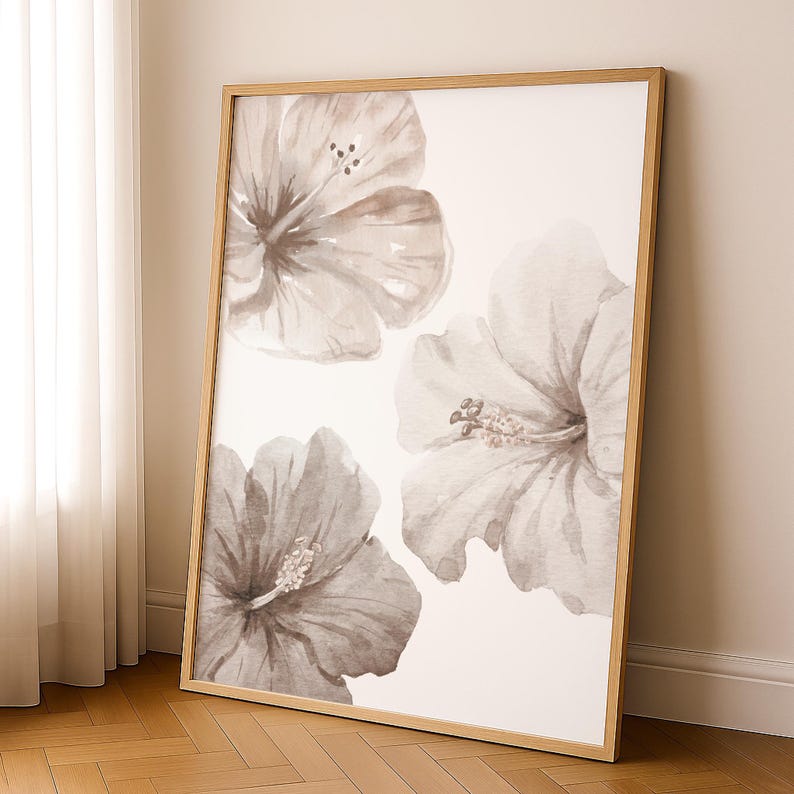 May include: A framed wall art print featuring three large watercolor hibiscus flowers in shades of gray and brown against a white background. The artwork is encased in a light-colored wooden frame, leaning against a wall.