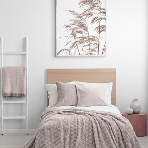 May include: A light pink plush throw blanket with a diamond pattern is draped over a bed with a wooden headboard. Two pillows are on the bed, one with a geometric pattern and the other a solid color. A framed print of dried grasses is on the wall above the bed.