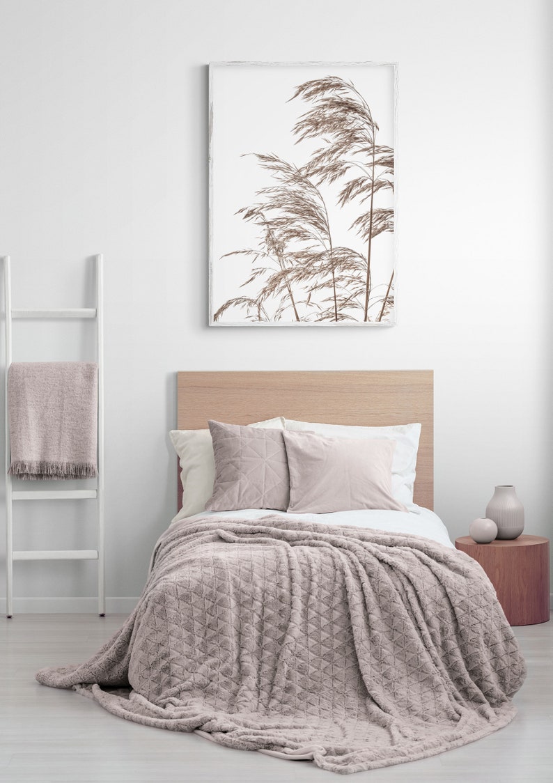 May include: A light pink plush throw blanket with a diamond pattern is draped over a bed with a wooden headboard. Two pillows are on the bed, one with a geometric pattern and the other a solid color. A framed print of dried grasses is on the wall above the bed.