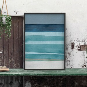 May include: A framed abstract watercolor painting with horizontal blue and teal stripes on a white background. The painting is hanging on a wall with a wooden door and a green table in front of it.