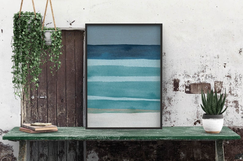 May include: A framed abstract watercolor painting with horizontal blue and teal stripes on a white background. The painting is hanging on a wall with a wooden door and a green table in front of it.