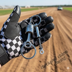 Puede incluir: Dos llaveros plateados, uno con forma de pistón de motor y otro de amortiguador, se sostienen sobre un fondo de pista de tierra. Los llaveros están unidos a un anillo grande. El texto "checkered charm racing" es visible.