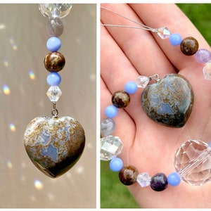 May include: A heart-shaped pendant made of brown and blue agate with a silver chain. The pendant is part of a beaded necklace with clear crystal, amethyst, and blue agate beads.