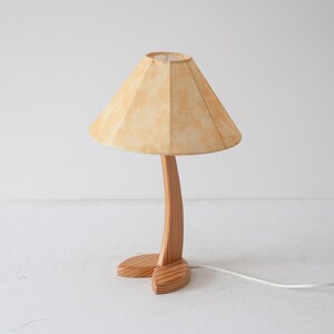 May include: A wooden table lamp with a light brown fabric shade. The lamp base is shaped like a stylised tree trunk with two feet.