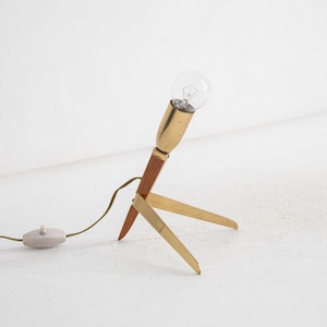 May include: A retro table lamp with a brass and wood construction. The lamp has a clear light bulb, a brown wooden stem, and three brass legs. It includes a cable with an on/off switch.
