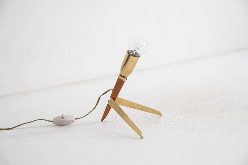 May include: A retro table lamp with a brass and wood construction. The lamp has a clear light bulb, a brown wooden stem, and three brass legs. It includes a cable with an on/off switch.