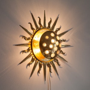 以下が含まれることがあります： 中央に三日月形の月がある太陽の形をした金色の金属製の壁掛け照明。月は質感のある表面で、小さな透明なガラス球で囲まれています。