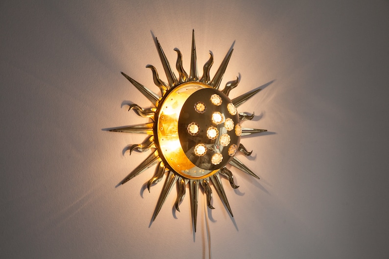 以下が含まれることがあります： 中央に三日月形の月がある太陽の形をした金色の金属製の壁掛け照明。月は質感のある表面で、小さな透明なガラス球で囲まれています。