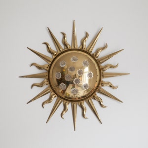 以下が含まれることがあります： 丸い中心と質感のある表面を持つ、金色のサンバーストの壁飾り。サンバーストの中心は、小さな透明なクリスタルで飾られています。