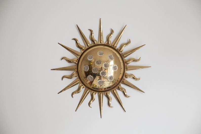 以下が含まれることがあります： 丸い中心と質感のある表面を持つ、金色のサンバーストの壁飾り。サンバーストの中心は、小さな透明なクリスタルで飾られています。