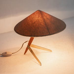 May include: A mid-century table lamp with a conical, brown lampshade. The lamp has a wooden base with three legs and a red support. The lamp is turned on, and the cord has a switch.